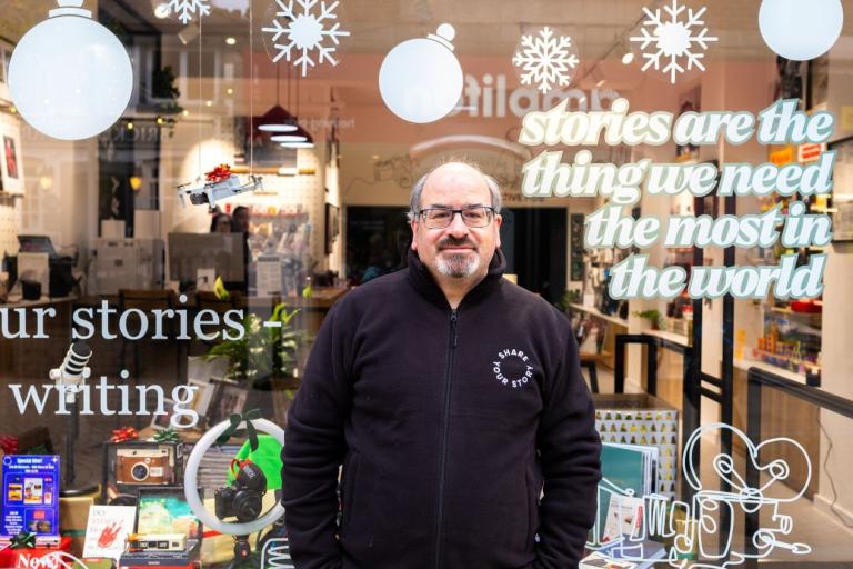 Photo shows the storefront of “Share Your Story” in Altrincham, featuring staff member Simon standing in front of the glass window.