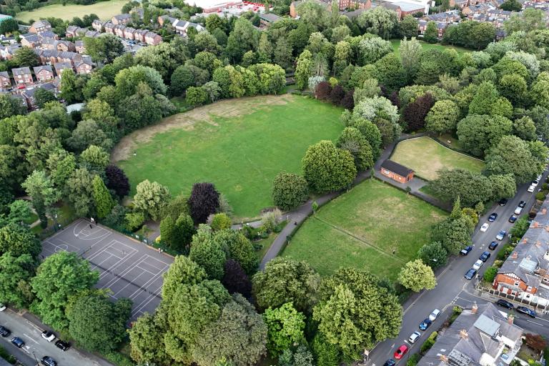 Aerial view of Stamford Park showing a large grassy field surrounded by dense trees, with a sports court, smaller lawn areas, and nearby residential streets lined with houses and parked cars.
