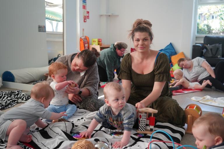 Babies and adults sitting and playing together on mats inside a bright, welcoming room with toys and blankets.