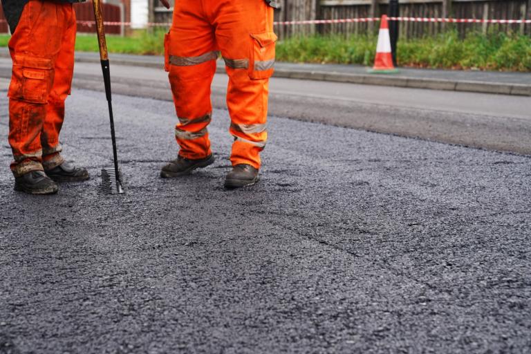 Two road workers in bright orange safety gear resurfacing a street with fresh tarmac.