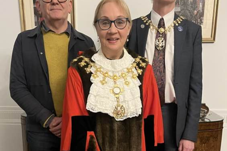 Three people stand indoors, with one in front wearing ceremonial mayoral robes and chains, and two others behind dressed in smart clothing, posing for a formal photograph in an office setting.