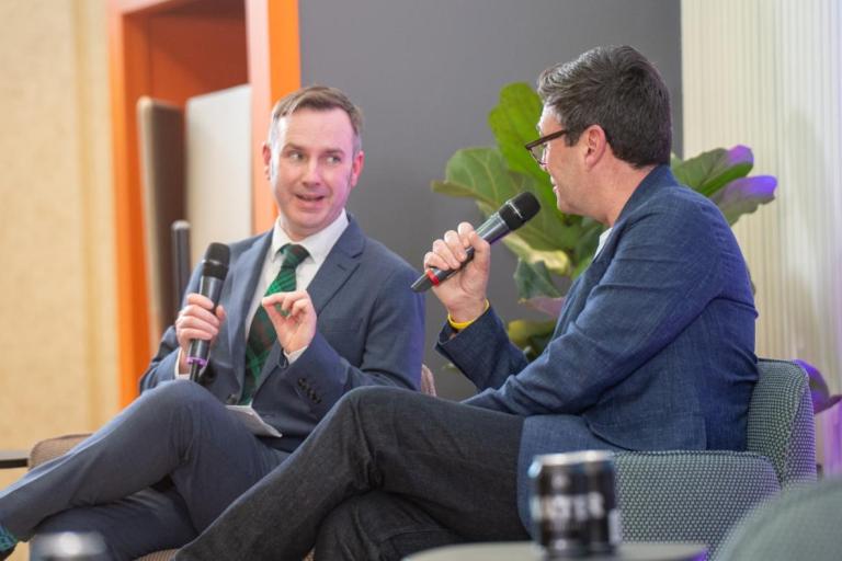 Two people sit on stage-facing chairs, each holding a microphone while engaged in conversation. 