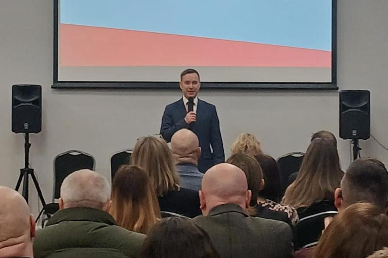 Cllr Tom Ross stands at the front of a conference room holding a microphone, addressing an audience seated in rows