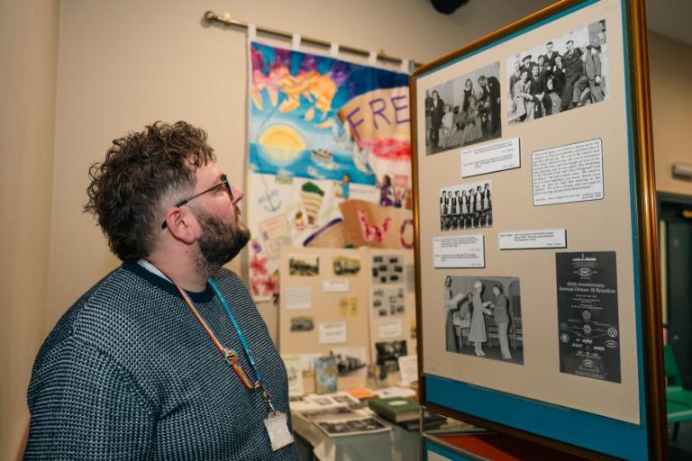 A man stands indoors looking at a display board filled with historical black‑and‑white photographs and printed captions.