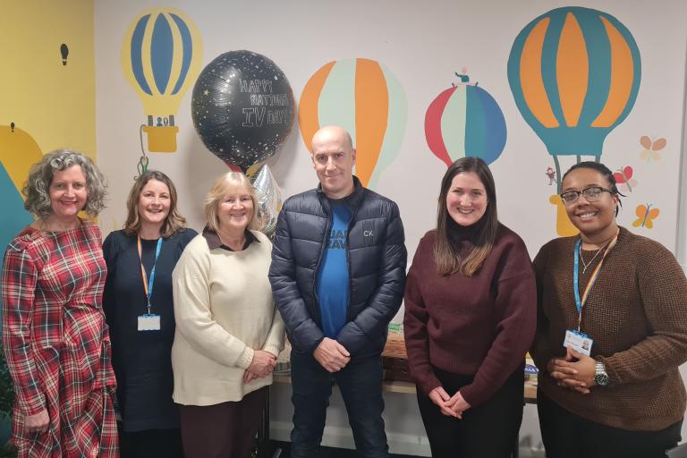 A group of six adults stand together in a brightly decorated room with colourful hot‑air balloon wall art and birthday balloons behind them.