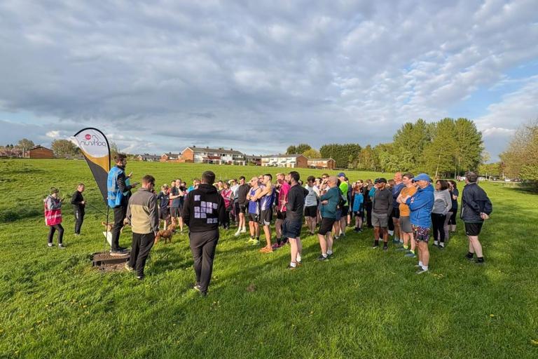 A large group of people gathered on a grassy field for a community running event, listening to a speaker standing on a small platform beside a flag banner. 