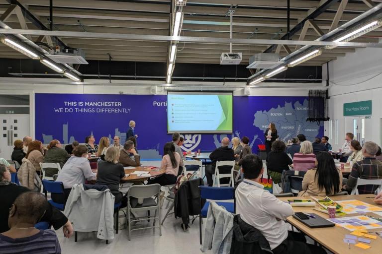 People seated at round tables in a large indoor event space watch a presentation projected on a screen, while two presenters stand at the front