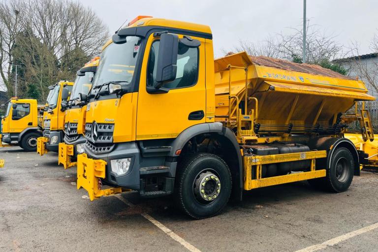Row of bright yellow grit-spreading lorries parked in a lot on an overcast day