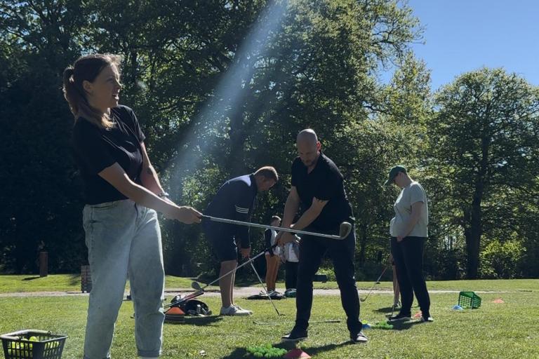 People practicing golf swings on a grassy driving range under bright sunlight, with several baskets of golf balls on the ground and a line of trees in the background.