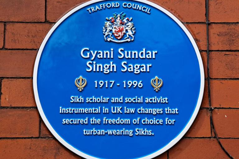 Blue circular commemorative plaque on a red brick wall reading “Trafford Council” with a crest above the name Gyani Sundar Singh Sagar (1917–1996). The plaque notes that he was a Sikh scholar and social activist instrumental in UK law changes securing freedom of choice for turban-wearing Sikhs.