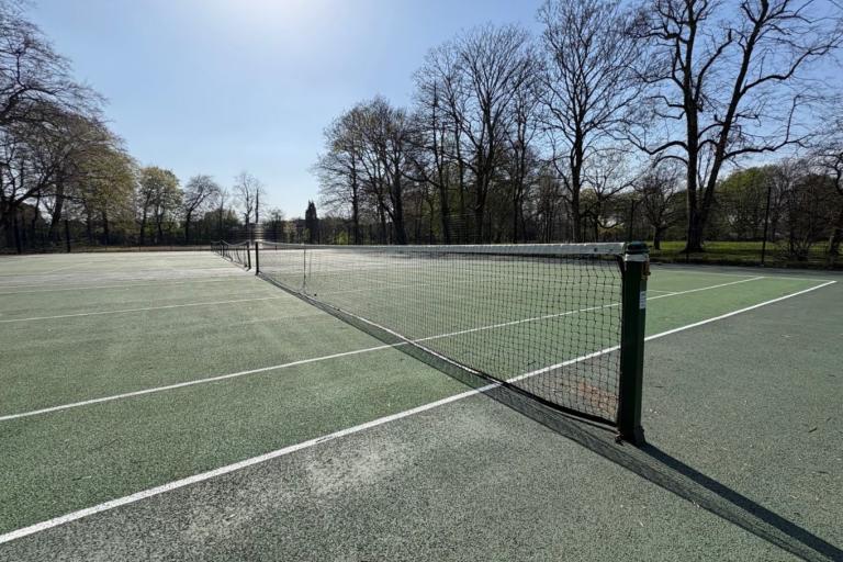 Empty outdoor tennis courts in Longford Park, viewed along the net stretching across the middle. The green hard surfaces are marked with white lines, and tall leafless trees line the background under a clear blue sky.
