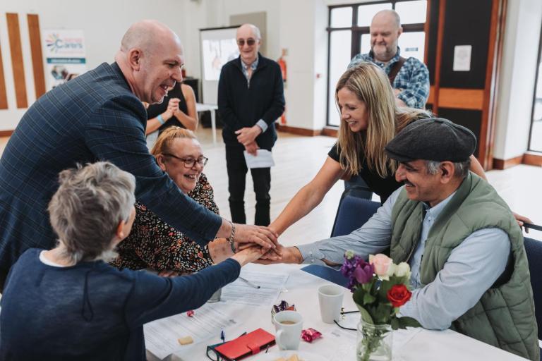 A group of adults gathered around a table indoors place their hands together in the center as part of a group activity. 