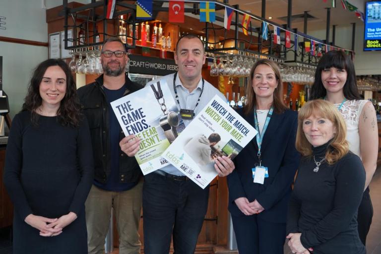Group of six adults in a bar holding domestic abuse awareness posters, with international flags and glassware visible behind them. from left to right: Cllr Keleigh Glenton, Executive Member for Community Safety and Civil Contingencies; John Lloyd, GMP Licensing Officer for Trafford; Ben Plunkett, Manager of The Bishop Blaize Wetherspoons; Nicola Walton, Domestic Abuse Co-ordinator, Trafford Council; Cllr Jo Harding, Deputy Mayor of Trafford and Executive Lead for Violence Against Women and Girls; Samantha F