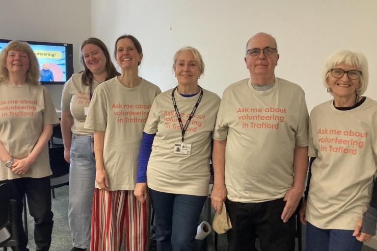 Six people stand side by side indoors wearing matching beige T‑shirts with the printed slogan “Ask me about volunteering in Trafford.” The group faces the camera in a room with chairs and a screen in the background displaying a volunteering-themed slide.