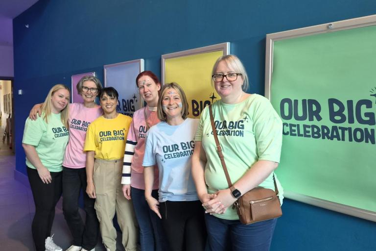 A group of seven people standing together indoors against a blue wall, wearing pastel T-shirts that read “Our Big Celebration,” with colorful framed panels behind them.