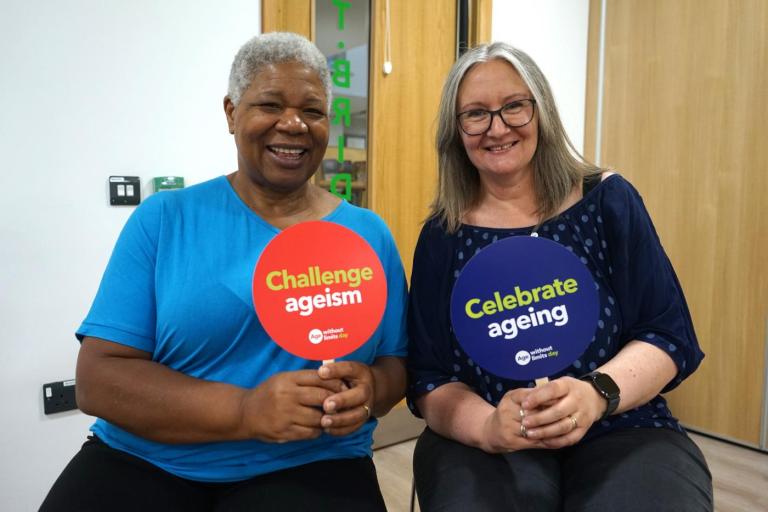 Two people seated indoors hold circular signs for Age Without Limits Day. One sign is red and reads “Challenge ageism,” and the other is purple and reads “Celebrate ageing.” The scene is in a bright room with wooden doors in the background.