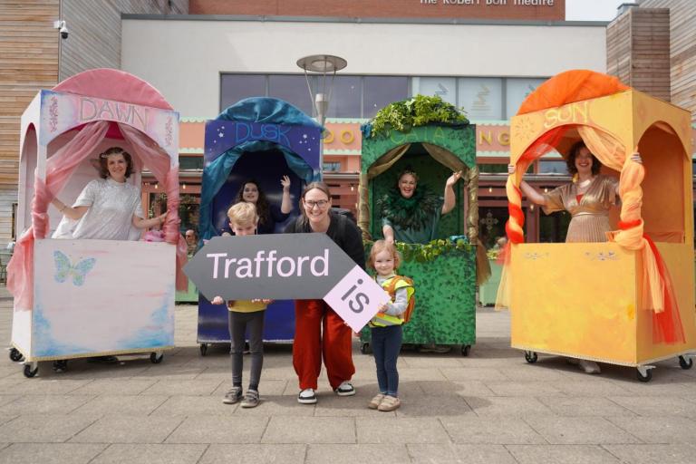 A group of people outside the Waterside Arts centre in Sale. In the background, four performers stand in colorful, wheeled booths labeled "Dawn," "Dusk," "Gaia," and "Sun." In the foreground, two adults and two children smile while holding a large, arrow-shaped sign that reads "Trafford Is".