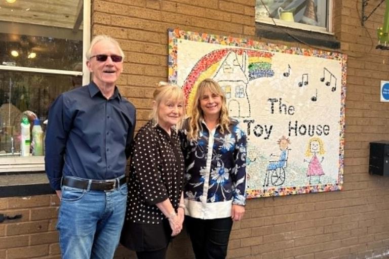 Three adults stand outside a brick building beside a large decorative sign reading “The Toy House.” The sign features a rainbow, musical notes, a house illustration, and drawings of children. A window is visible on the left side of the building, and the group is posed in front of the sign.