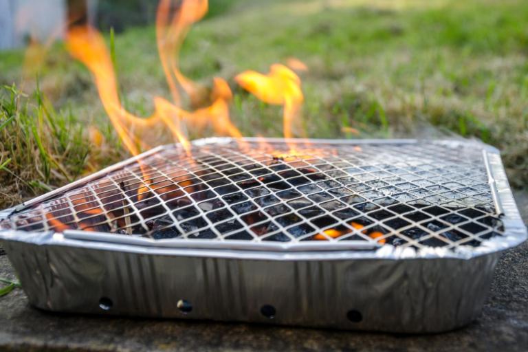 Disposable foil barbecue tray with a wire grill and glowing charcoal, producing visible flames outdoors on a stone surface beside a grassy lawn.