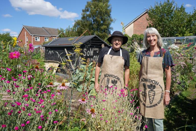 Two people wearing aprons with the Bowdon Allotment and Garden Society logo stand among colourful flowers and vegetable beds at an allotment garden. Behind them are a black garden shed, greenhouses and trees.