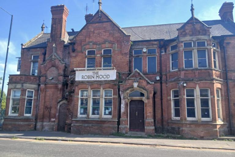 Front view of a large, disused red-brick Victorian building with Gothic architectural details, chimneys, bay windows, and a central entrance.