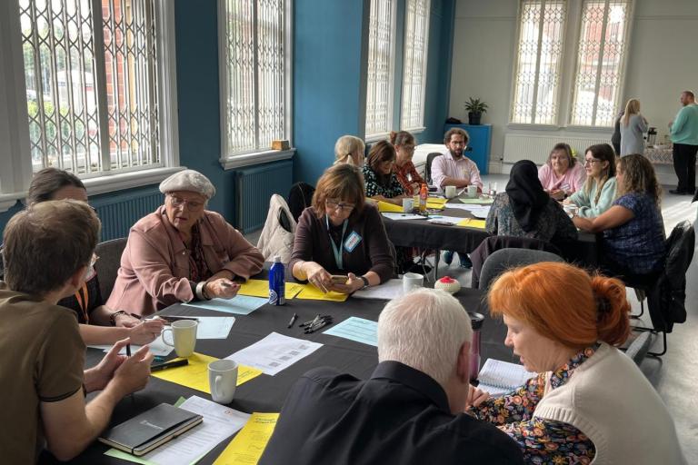 A community workshop taking place in a bright meeting room with large blue walls and tall windows fitted with security grilles. Around a dozen participants are seated at round tables covered with black tablecloths, discussing and completing worksheets.